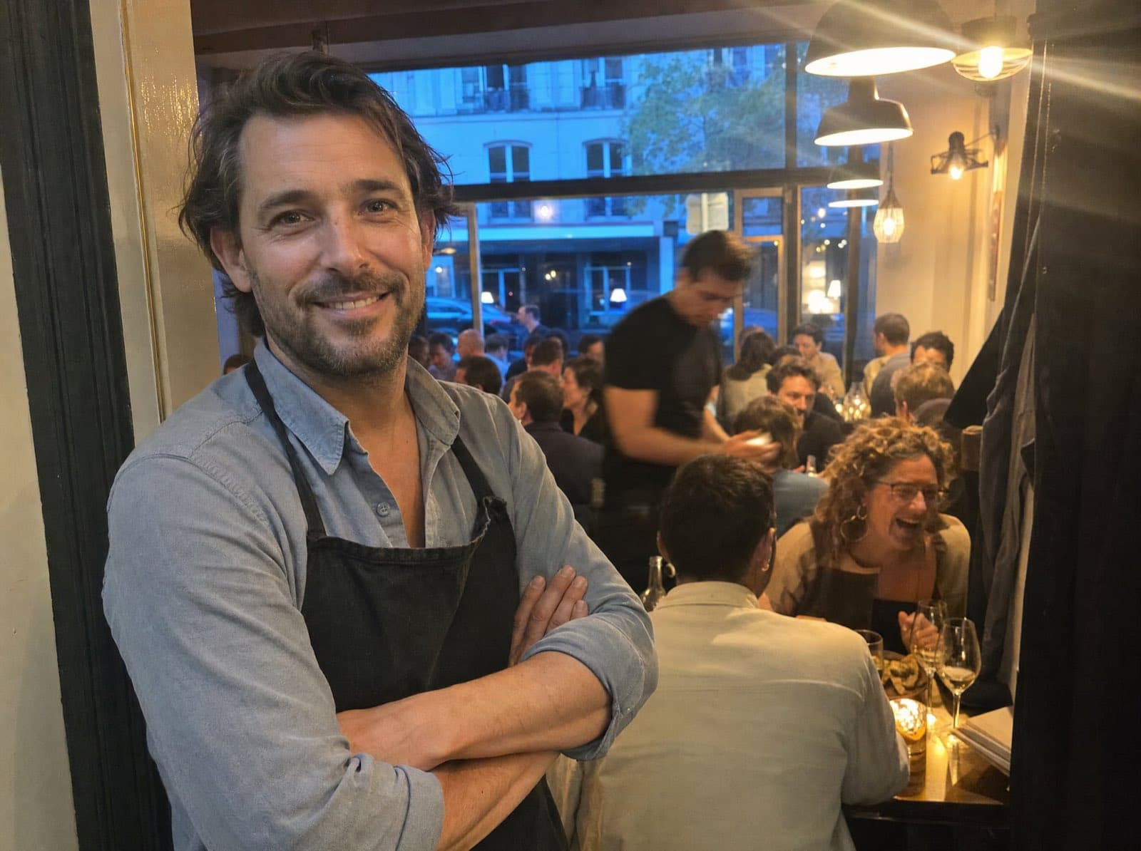 A restaurant owner in a dark apron stands smiling in the doorway of his own bistro, with a full dining room behind him.